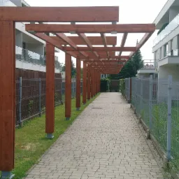 Pergola z belek drewnianych nad ścieżką z kostki brukowej, widok wzdłuż, z metalowymi ogrodzeniami po bokach, budynki mieszkalne w tle.