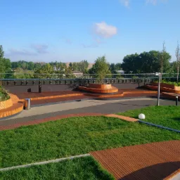 Widok z góry na nowoczesny taras z drewnianymi elementami, trawnikiem, roślinnością i oświetleniem, z balustradą w tle i widokiem na okolicę.