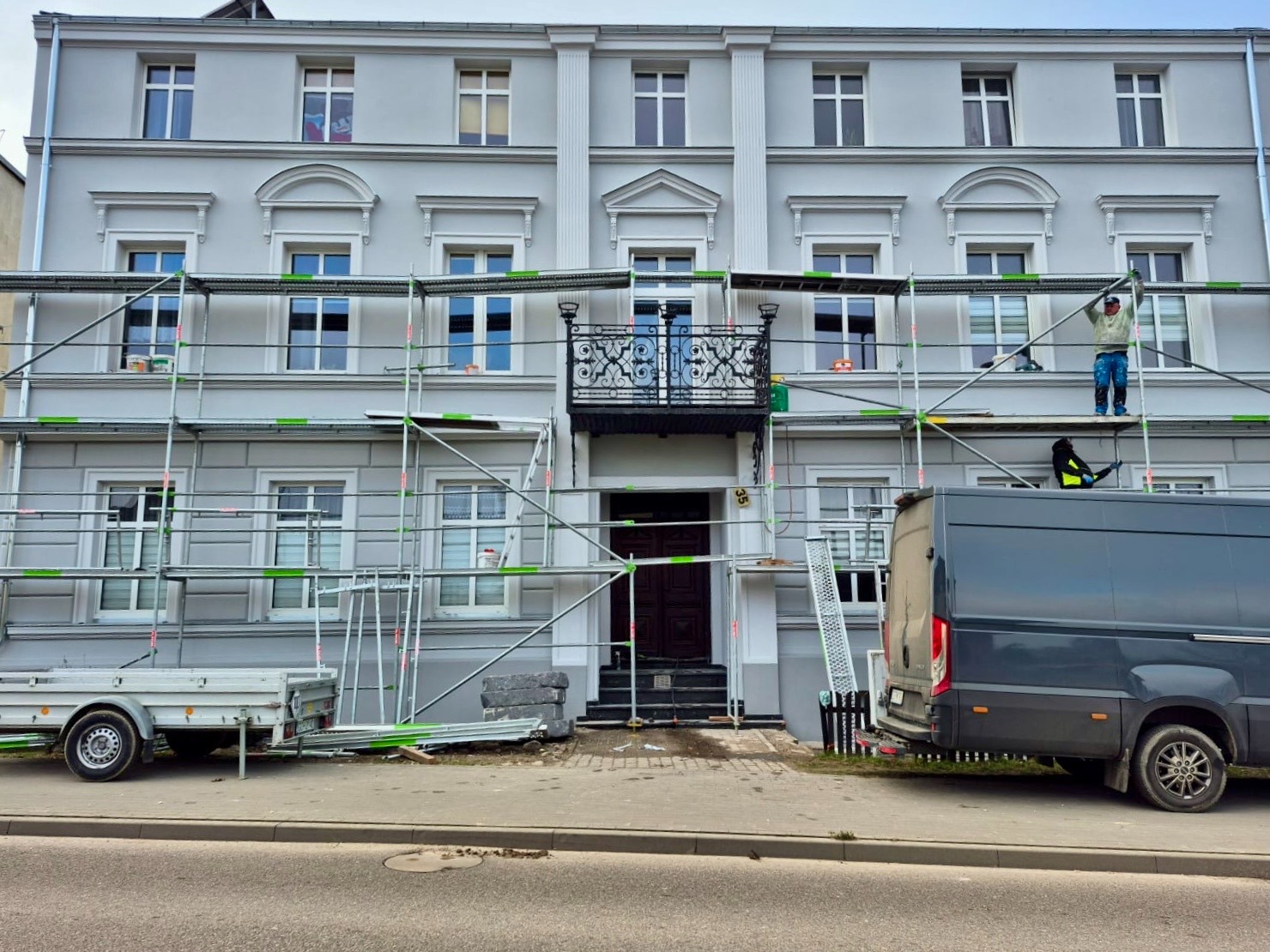 Dwóch pracowników na rusztowaniu podczas odnawiania elewacji kamienicy w odcieniach szarości, z zaparkowanym vanem i przyczepą na pierwszym planie.