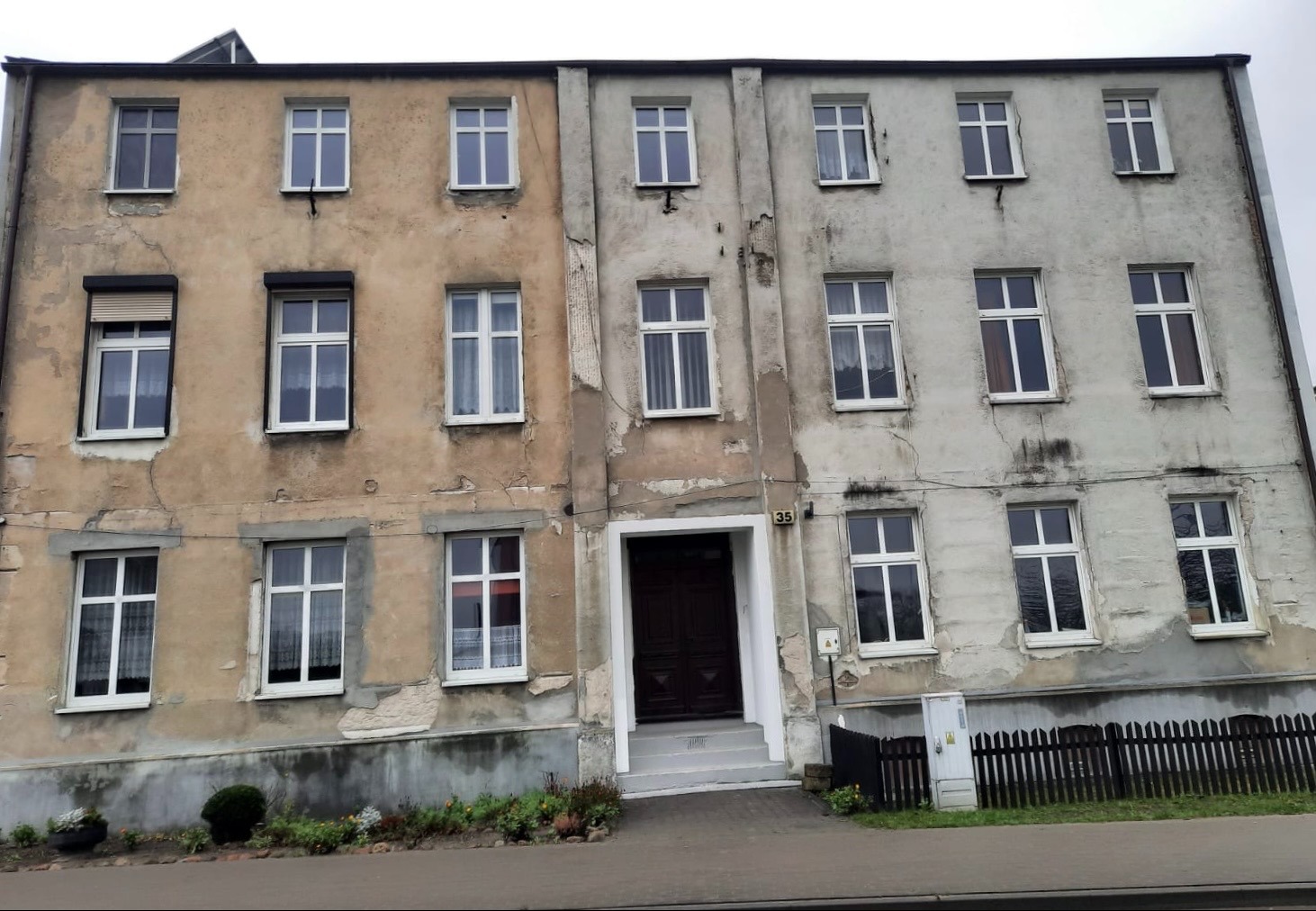 Dwukondygnacyjny budynek przed remontem, z jedną częścią pomalowaną na beżowo, a drugą w surowym, szarym betonie, ukazujący różnicę w stanie elewacji.