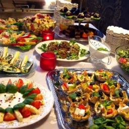 Bogato zastawiony stół z różnorodnymi przekąskami, sałatkami i deserami, w tym mini tartaletki z warzywami, koreczki i sałatka caprese, oświetlony ciepłym światłem.