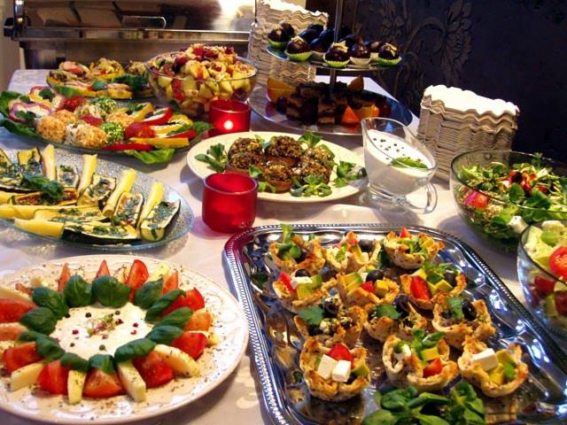 Bogato zastawiony stół z różnorodnymi przekąskami, sałatkami i deserami, w tym mini tartaletki z warzywami, koreczki i sałatka caprese, oświetlony ciepłym światłem.