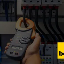 Pomiar prądu cęgowym miernikiem na tle rozdzielnicy elektrycznej z bezpiecznikami i przewodami.