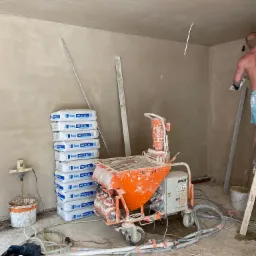 Wnętrze w trakcie prac wykończeniowych, ściany pokryte świeżym tynkiem, widoczna pomarańczowa maszyna do tynkowania PFT z zabrudzeniami, ułożone worki z zaprawą Knauf MP 75 SL oraz pracownik...