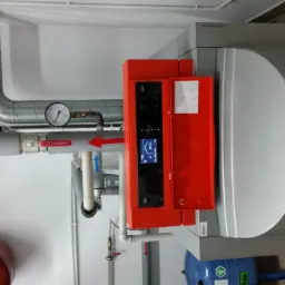 Nowoczesny piec gazowy z czerwonym panelem sterowania, widoczne rury instalacji grzewczej z manometrem, zbiornik wyrównawczy w tle.