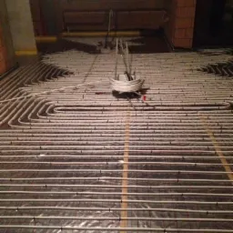 Rozłożone rury ogrzewania podłogowego na folii z siatką, widoczny zwój rur i fragment muru z cegły