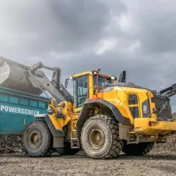 Żółta ładowarka Volvo H150H nabiera urobek do przesiewacza Powerscreen na placu budowy, w tle hałda kamieni i pochmurne niebo.