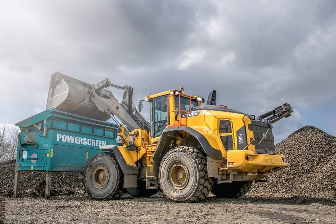 Żółta ładowarka Volvo H150H nabiera urobek do przesiewacza Powerscreen na placu budowy, w tle hałda kamieni i pochmurne niebo.