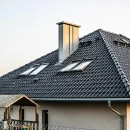 Ciemna dachówka ceramiczna na skośnym dachu z trzema oknami dachowymi i metalowym kominem, widok z dołu na fragment budynku z drewnianą werandą.