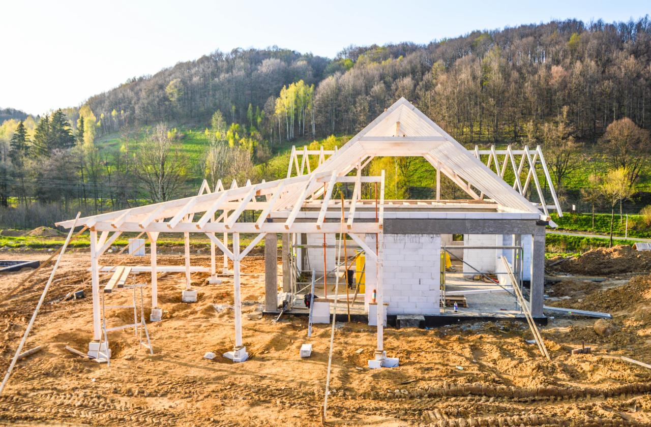 Budowa domu w Zagórzu Śląskim: surowy szkielet konstrukcji z białych bloczków i drewnianej więźby dachowej na tle zalesionego wzgórza w słoneczny dzień.