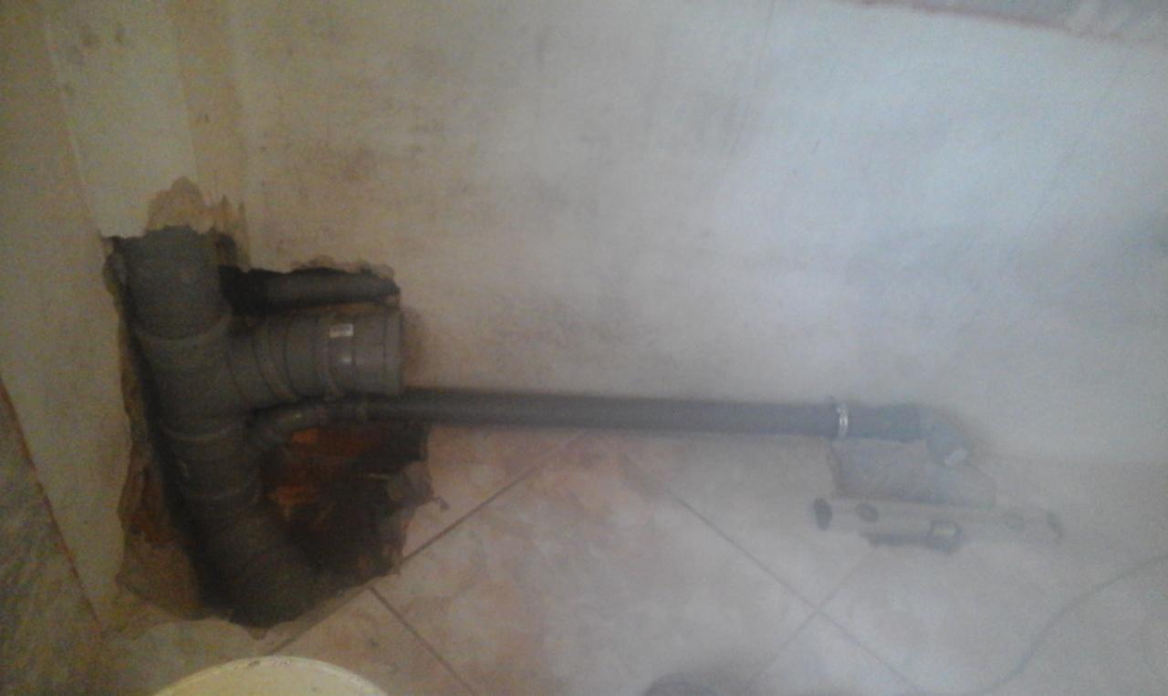 Szare rury kanalizacyjne wystające ze ściany, częściowo zamurowane, widoczne płytki na podłodze i niedokładne wykończenie ściany.