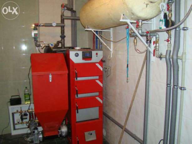 Czerwony piec centralnego ogrzewania z zasobnikiem na paliwo, widoczne rury instalacji grzewczej i zbiornik akumulacyjny, całość w pomieszczeniu z wykafelkowanymi ścianami.