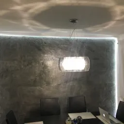 Ściana w jadalni wykończona szarą okładziną przypominającą kamień, oświetlona taśmą LED wzdłuż krawędzi sufitu, z nowoczesną lampą wiszącą i czarnymi krzesłami przy stole.