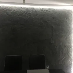 Ściana wykończona szarą płytą imitującą kamień z podświetleniem LED, widoczne dwa krzesła i fragment zasłony.