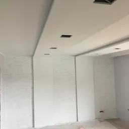 Wnętrze pokoju w trakcie wykańczania; fragment ściany z dekoracyjną fakturą, gładkie panele ścienne, sufit z otworami na oświetlenie, podłoga częściowo zabezpieczona.