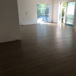 Wnętrze po remoncie z ułożonymi panelami podłogowymi w odcieniach brązu, widoczne białe ściany, okna i wystające kable ze stropu.