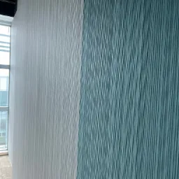 Ściana z tapetą w dwóch odcieniach: jasnoszarym i turkusowym, z wyraźną fakturą pionowych pasków, widoczne okno z żaluzjami w tle.