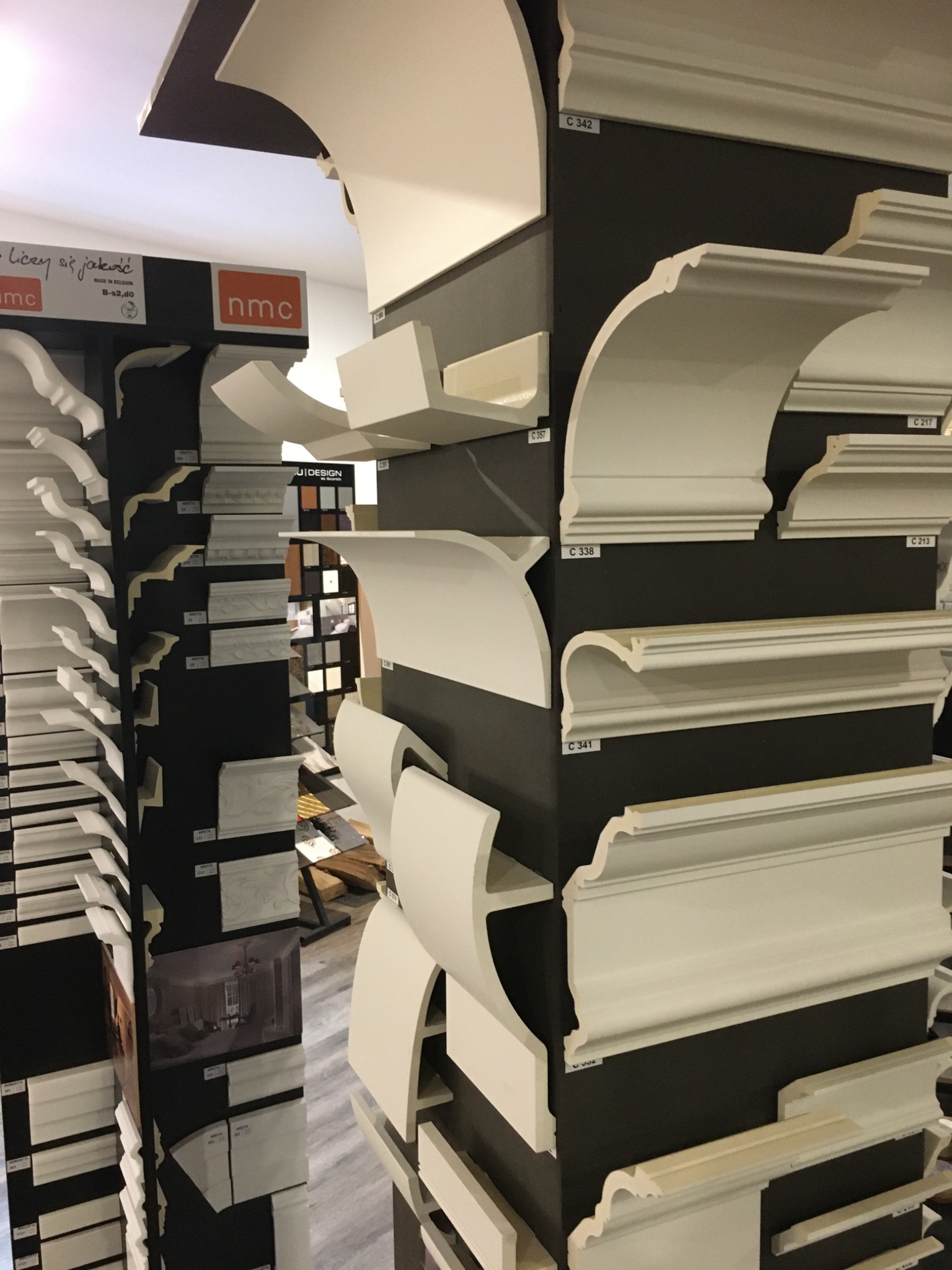 Ekspozycja różnorodnych, białych listew dekoracyjnych i sztukaterii, przytwierdzonych do ciemnych, pionowych paneli w sklepie. Widoczne numery katalogowe przy każdym modelu.