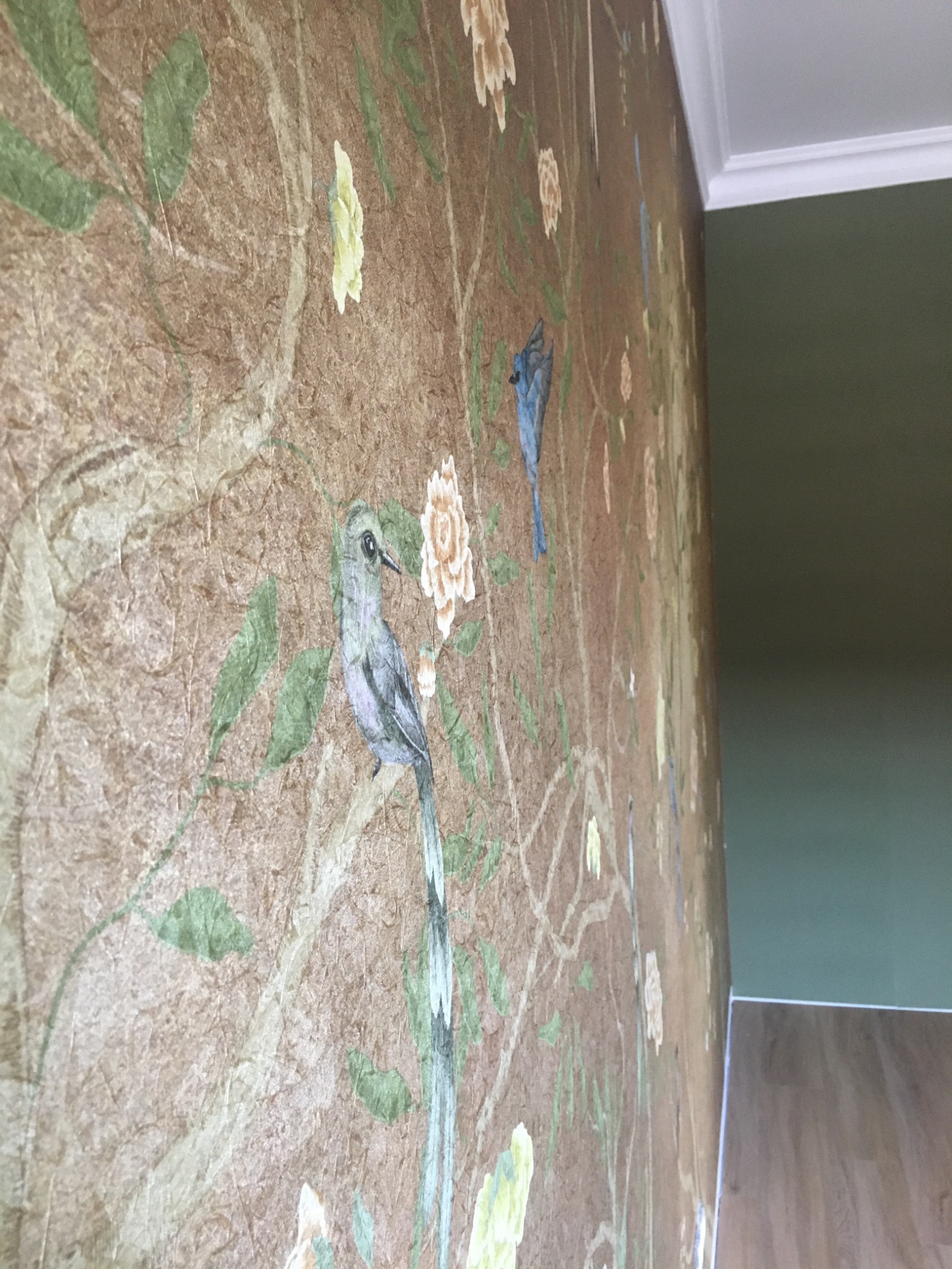 Beżowa ściana z wzorem ptaków i kwiatów, widoczna pod kątem, z fragmentem zielonej ściany i białego sufitu w tle.