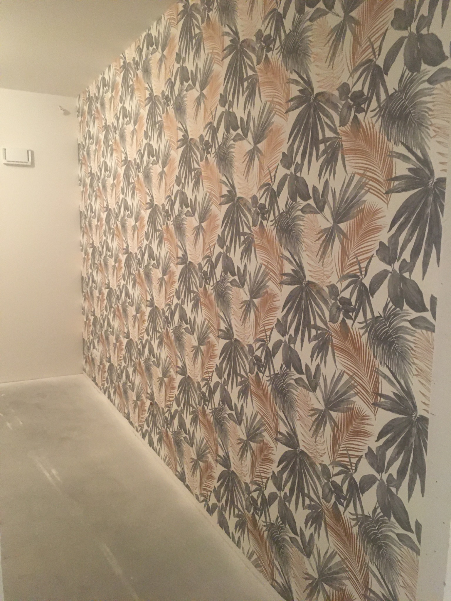 Ściana po nałożeniu tapety z motywem tropikalnych liści w odcieniach szarości i brązu, widok z perspektywy narożnika pomieszczenia.