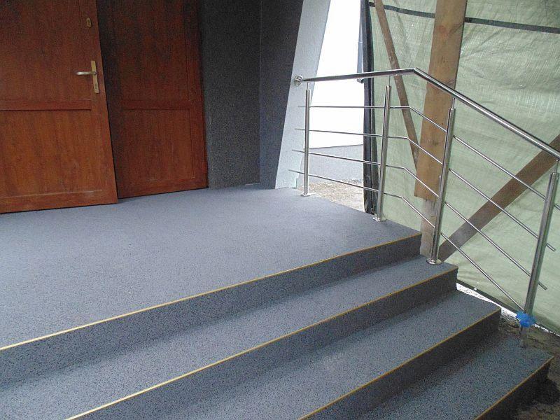 Szare schody zewnętrzne pokryte żywicą z metalową balustradą i drewnianymi drzwiami wejściowymi z boku.