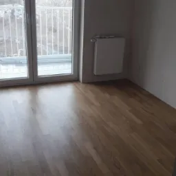 Świeżo położona, błyszcząca podłoga z paneli w pustym pokoju z balkonem i grzejnikiem na ścianie.