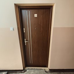 Brązowe drzwi wejściowe z numerem 28 i srebrnymi paskami, zamontowane w bloku. Widoczne ślady po montażu i niedomalowane fragmenty ściany.