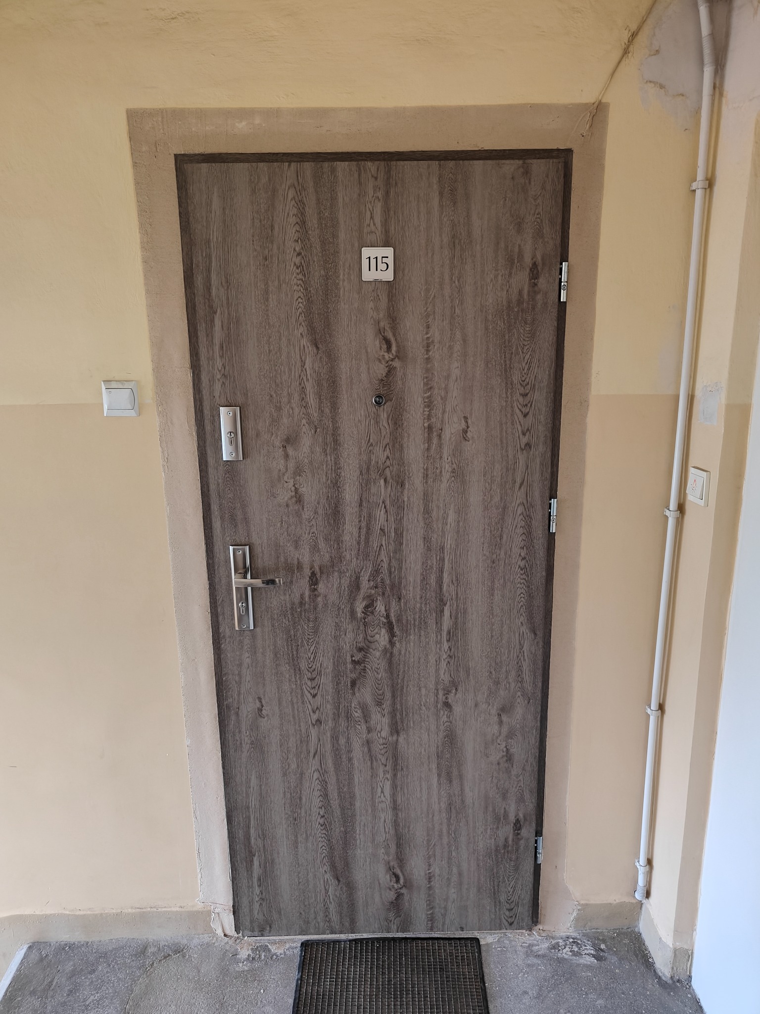 Drzwi wejściowe do mieszkania z numerem 115, w odcieniach szarości z drewnopodobnym wzorem, zamontowane w beżowej futrynie i ścianie. Widoczny zamek i klamka.