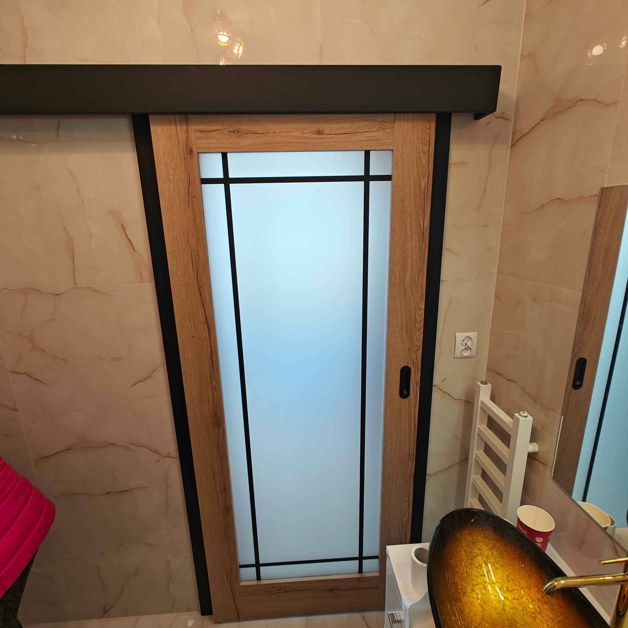 Zamontowane przesuwne drzwi łazienkowe z drewnianą ramą i matowym szkłem, czarne detale, widok na umywalkę i grzejnik drabinkowy, marmurowe płytki na ścianach.