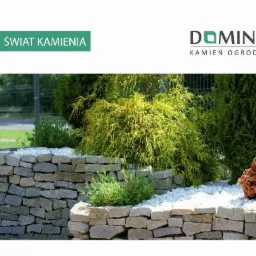 Ogródek skalny z kamienną obudową, wypełniony białym grysem i ozdobnymi iglakami oraz czerwoną skałą w tle, widoczny fragment ogrodzenia.