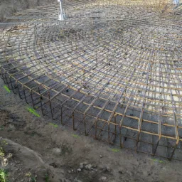 Zbrojenie fundamentu w wykopie, widoczne stalowe pręty ułożone w siatkę z elementami dystansowymi, ziemia wokół z zielonymi oznaczeniami.