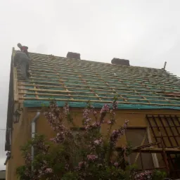 Montaż łat i kontrłat na dachu z widoczną membraną dachową Eurotop N, dwóch dekarzy na dachu, dom z kwitnącym krzewem na pierwszym planie, pochmurne niebo.