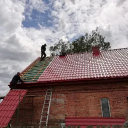 Prace dekarskie na dachu z czerwonej blachy trapezowej, widoczna więźba dachowa, trzech dekarzy na dachu, ceglany budynek z drewnianymi oknami w tle.