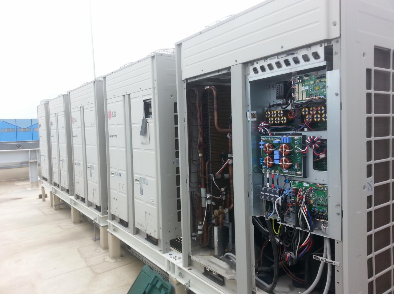Rząd białych jednostek zewnętrznych klimatyzacji LG Multi V na dachu budynku, z otwartym panelem ukazującym wewnętrzne okablowanie i komponenty elektryczne.
