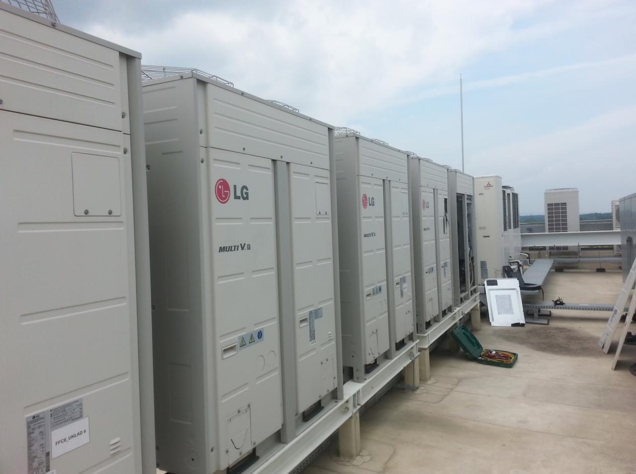 Rząd zewnętrznych jednostek klimatyzacyjnych LG Multi V na dachu budynku, widoczne narzędzia serwisowe i drabina.