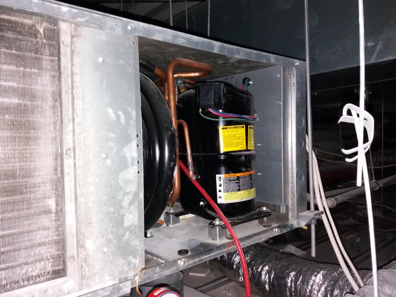 Wnętrze jednostki klimatyzacyjnej z widocznym kompresorem, miedzianymi rurkami, przewodami elektrycznymi i elementami metalowej obudowy, umieszczonej w przestrzeni technicznej z rurami i przewodami...