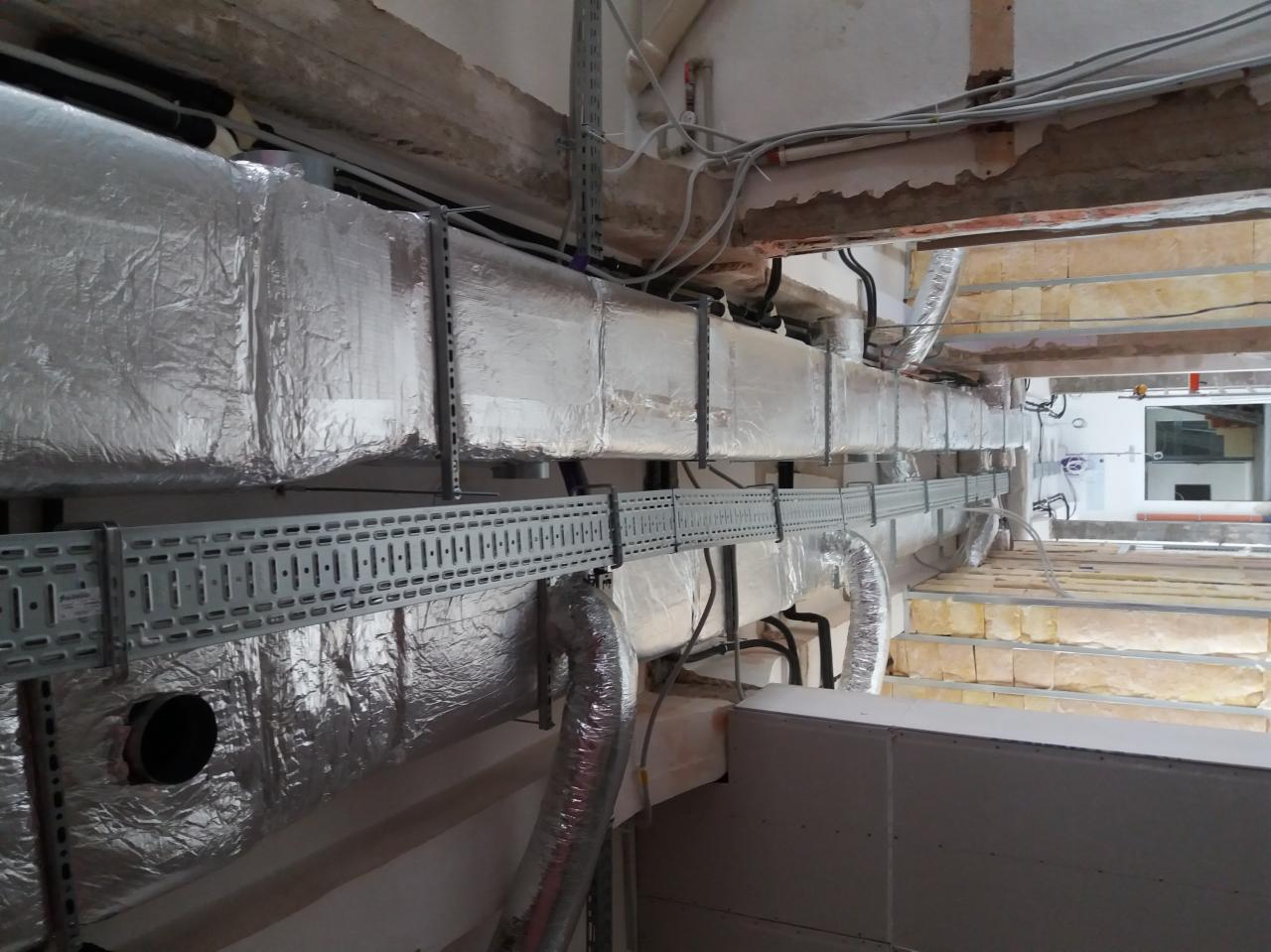Widok instalacji wentylacyjnej z izolowanymi przewodami w srebrnej folii, prowadzonej pod sufitem w surowym stanie. Widoczne metalowe wsporniki i koryta kablowe.