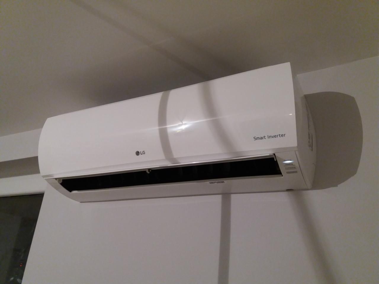 Biała jednostka klimatyzacji LG Smart Inverter zamontowana na ścianie w pomieszczeniu z widocznym oknem w tle.