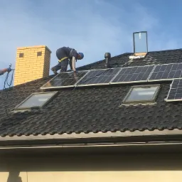 Montaż paneli słonecznych na dachu z dachówką karpiówką, widoczny komin z cegły klinkierowej i okno dachowe, dwóch monterów w kaskach zabezpieczonych linami.