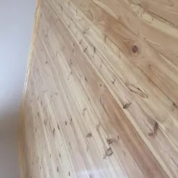 Jasne, lakierowane deski ułożone ukośnie na ścianie, widoczne sęki i naturalny wzór drewna, fragment białego sufitu w lewym górnym rogu.