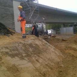 Pracownik w pomarańczowym kombinezonie i żółtym kasku stoi na ziemi obok wykopu, w tle mała koparka i konstrukcja mostu, na zboczu widoczne elementy stabilizujące grunt.