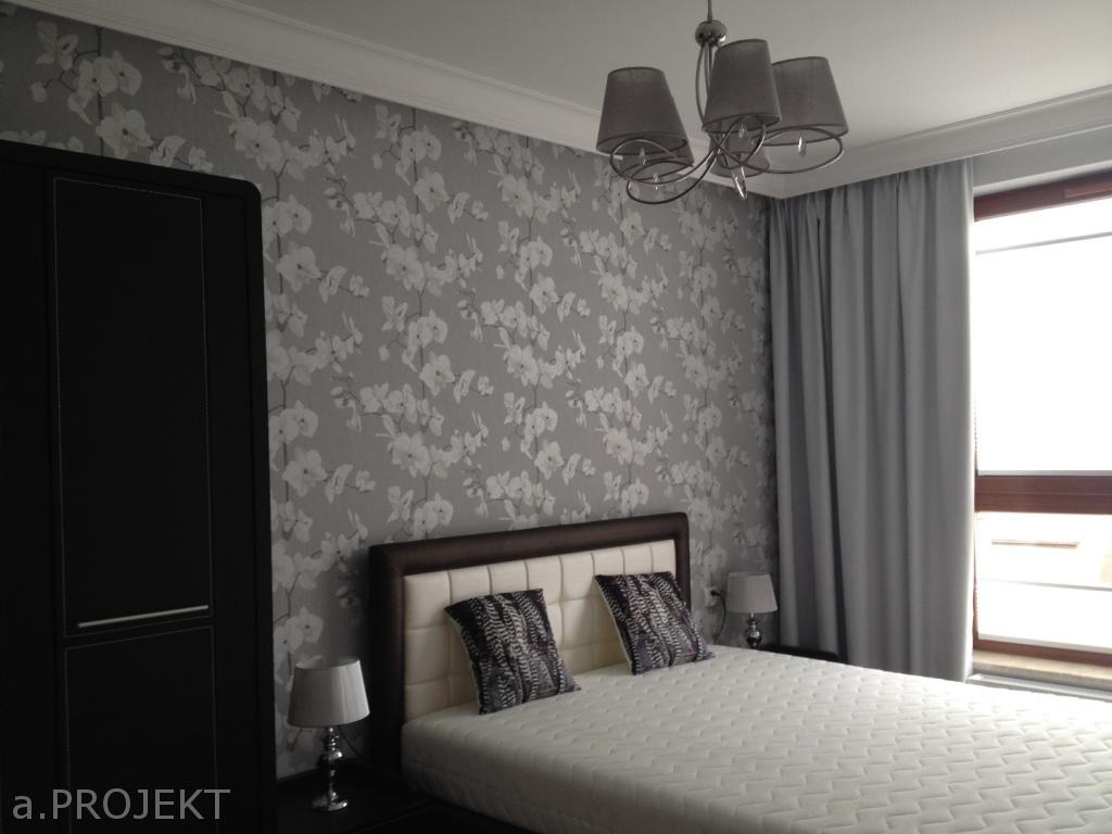 Sypialnia z tapetą w kwiaty, ciemną szafą i łóżkiem z pikowanym zagłówkiem, oświetlona lampą sufitową z szarymi abażurami.