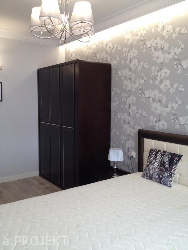 Sypialnia z ciemnobrązową szafą, łóżkiem z białym zagłówkiem i poduszką w etniczne wzory, tapeta w kwiaty i nowoczesna lampa sufitowa z abażurami.