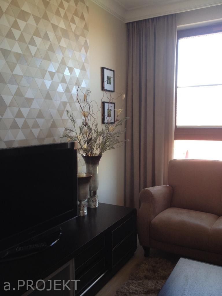 Salon z dekoracyjną ścianą w trójkąty, ciemną szafką RTV, dwoma wazonami z suchymi gałązkami oraz beżową sofą przy oknie z zasłonami.