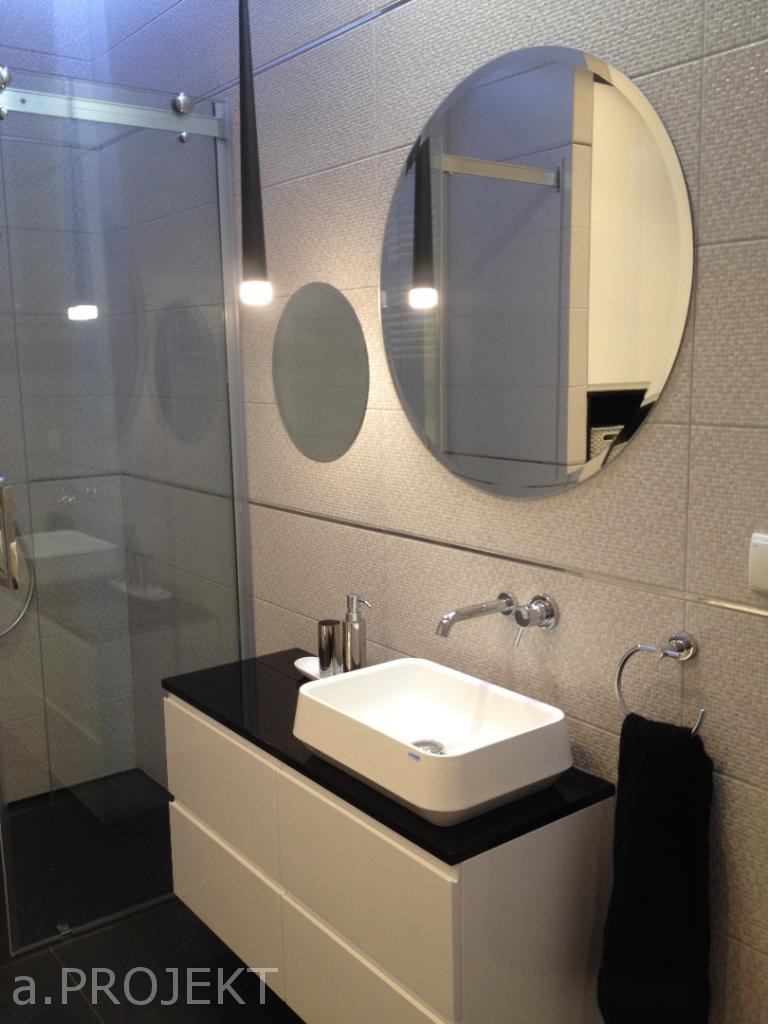 Nowoczesna łazienka z owalnym lustrem, podwieszaną szafką z białą umywalką nablatową i czarnym blatem oraz kabiną prysznicową ze szklanymi drzwiami przesuwnymi.