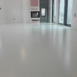 Biała, błyszcząca podłoga w nowym, jasnym pomieszczeniu z dużymi oknami balkonowymi. Widoczne ślady po taśmie malarskiej przy ścianach.