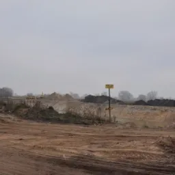 Rozległy teren po wykopie, z widocznymi hałdami ziemi i pracującą w oddali koparką, w tle zarośla i pochmurne niebo, żółta tablica ostrzegawcza na pierwszym planie.