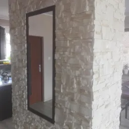 Ściana działowa wykończona dekoracyjnym kamieniem w odcieniach bieli i beżu, z wkomponowanym lustrem w czarnej ramie, odbijającym fragment wnętrza z drewnianymi drzwiami.