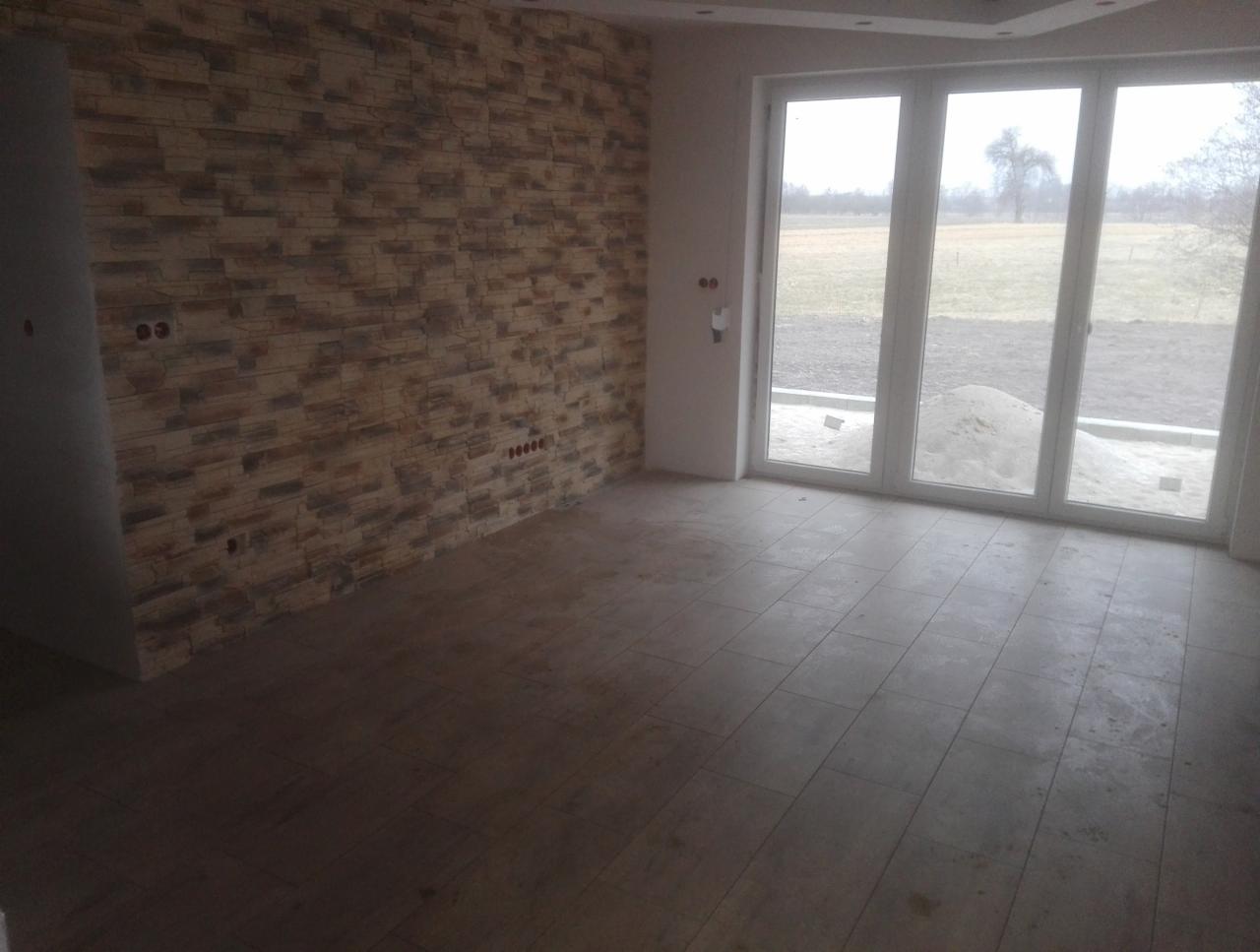 Wnętrze pokoju z wykończoną ścianą z dekoracyjnego kamienia, szarą podłogą z płytek i dużym, trzyskrzydłowym, białym oknem balkonowym z widokiem na pole.