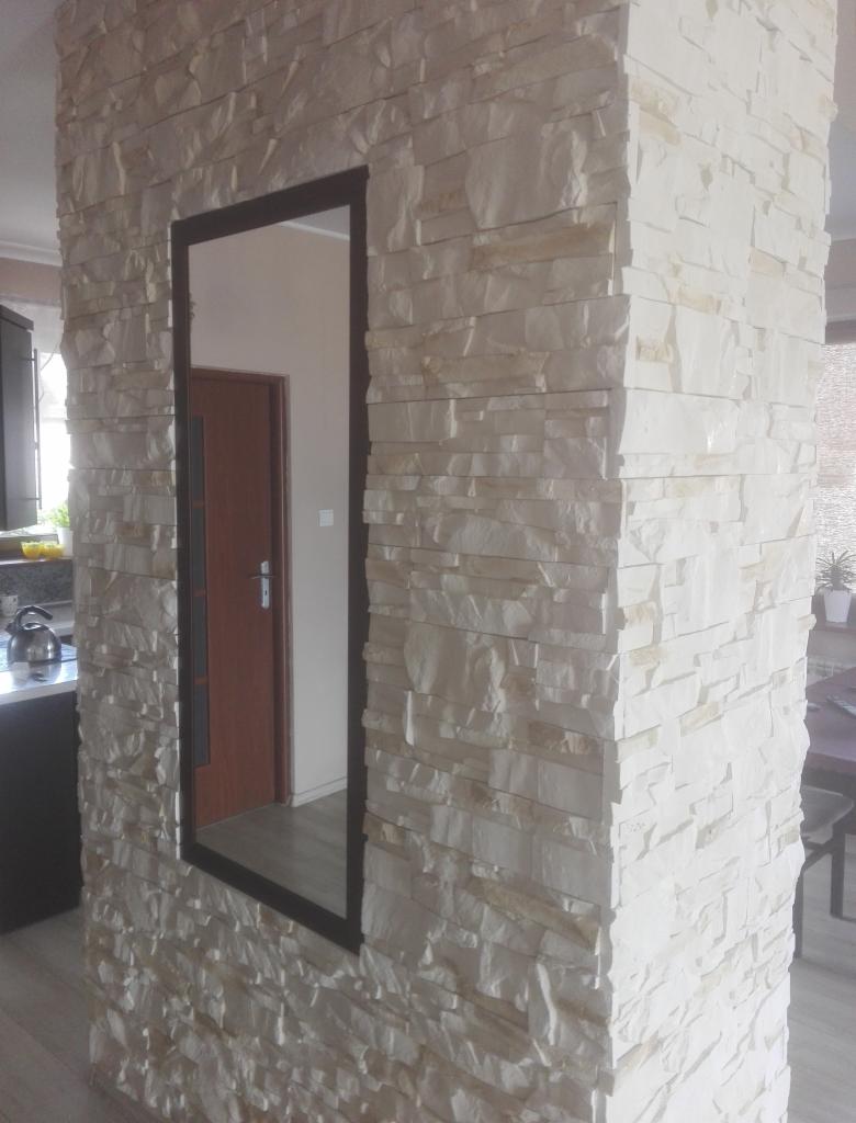 Ściana działowa wykończona dekoracyjnym kamieniem w odcieniach bieli i beżu, z wkomponowanym lustrem w czarnej ramie, odbijającym fragment wnętrza z drewnianymi drzwiami.